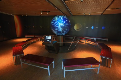 A planetarium room with a large globe centerpiece and benches arranged in a circle, featuring a dark wall with planets.
