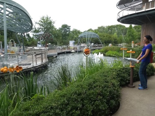 A person interacts with a water feature in a park, surrounded by greenery and a pond with fountains.