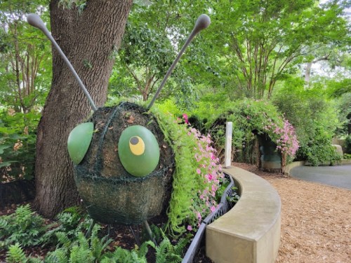 A whimsical topiary insect made of greenery and flowers, surrounded by lush plants in a garden setting.