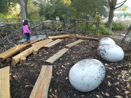 A child in a pink jacket climbs a wooden path near large, speckled concrete eggs in a natural play area.
