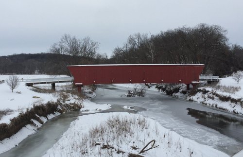 A red covered bridge spans a snowy landscape, with a frozen river and trees in the background.
