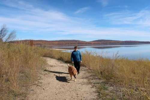 A person walks a golden retriever along a scenic lakeside path with tall grass and autumn foliage in the background.