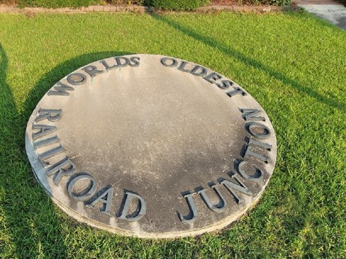 A circular stone plaque reading "World's Oldest Railroad Junction" on green grass.