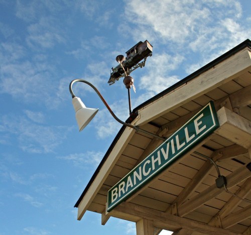 Sign for Branchville under a blue sky, featuring a lamp and a weather vane on a building's roof.