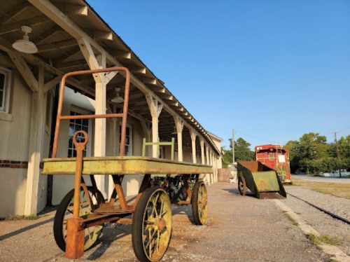 An old, rusty cart and a vintage vehicle near a building under a clear blue sky.