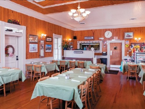 Cozy restaurant interior with wooden walls, green tablecloths, and a chandelier, featuring empty tables and a counter.