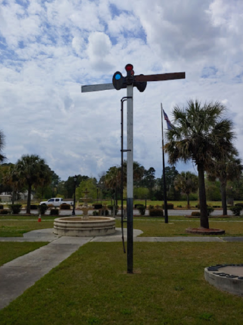 A railroad signal with blue and red lights stands in a grassy area, surrounded by palm trees and a cloudy sky.
