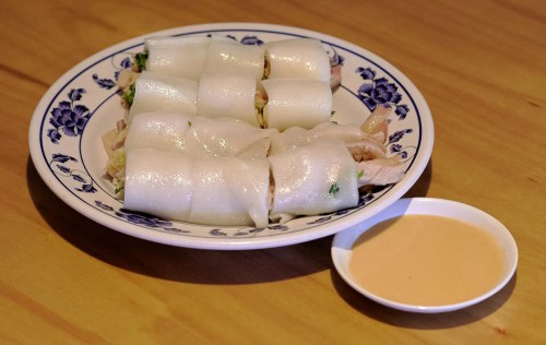 A plate of rice noodle rolls filled with chicken and herbs, served with a small bowl of dipping sauce.