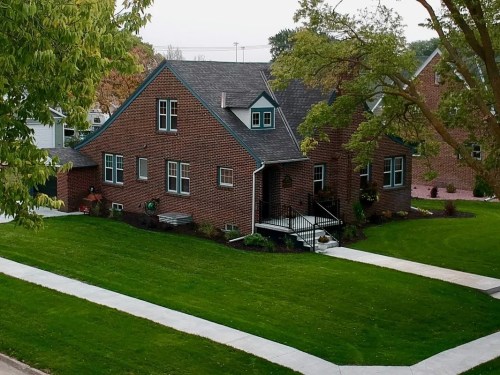 A brick house with green accents, surrounded by a well-maintained lawn and pathways, set in a residential area.