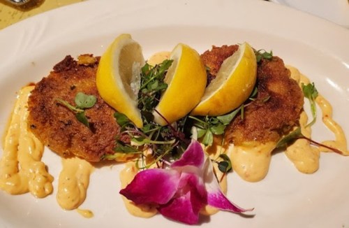 Three golden crab cakes topped with lemon slices and microgreens, drizzled with a creamy sauce and garnished with a flower.