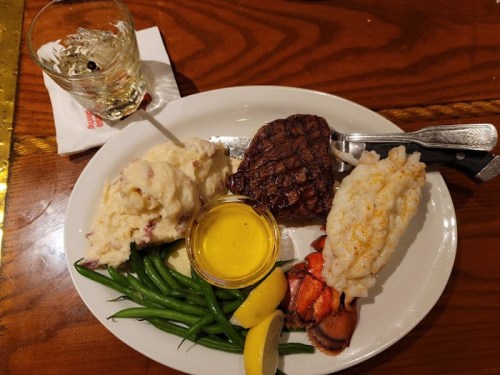 A plate featuring steak, lobster, mashed potatoes, green beans, and a glass of whiskey on the side.
