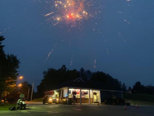 Fireworks illuminate the night sky above a small store, with a motorcycle parked nearby and trees in the background.