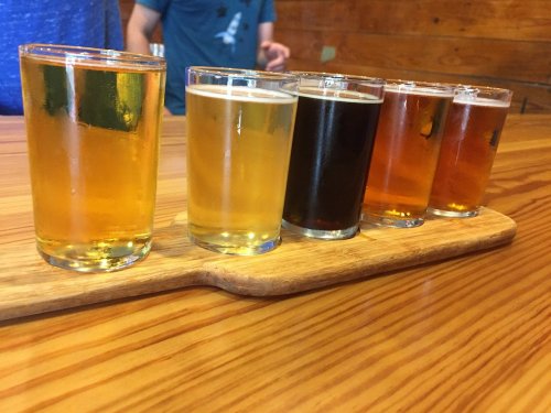 A wooden flight board holds five small glasses of different beers, ranging from light to dark, on a wooden table.