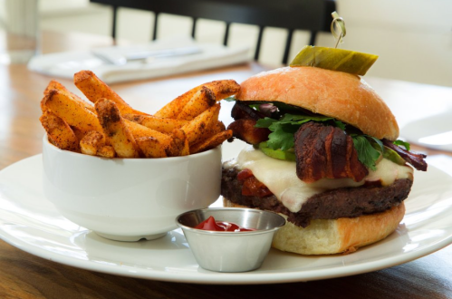 A juicy burger with bacon, cheese, and greens, served with a side of seasoned fries and a small cup of ketchup.