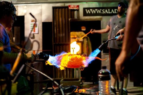 A glassblower shapes molten glass with flames, showcasing vibrant colors and intense heat in a workshop setting.