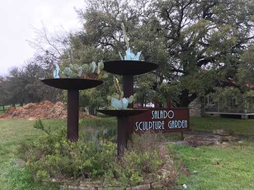 Sign for Salado Sculpture Garden featuring metal sculptures with cacti, surrounded by greenery and trees.