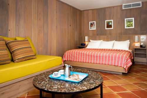 Cozy hotel room featuring a bed, yellow sofa, coffee table, and wooden walls with framed artwork.