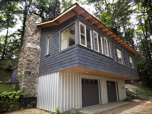 A modern two-story house with a stone chimney, large windows, and a metal garage door, surrounded by trees.