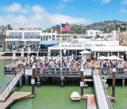 A bustling pier with people dining, an American flag flying, and waterfront buildings in the background.
