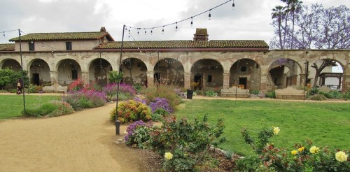 A historic building with arches surrounded by colorful flowers and greenery, under a cloudy sky with string lights.