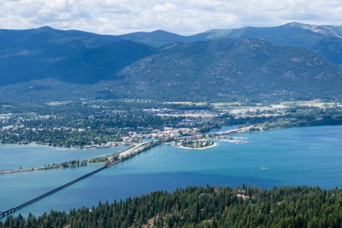 Aerial view of a town by a lake, surrounded by mountains and forests under a partly cloudy sky.