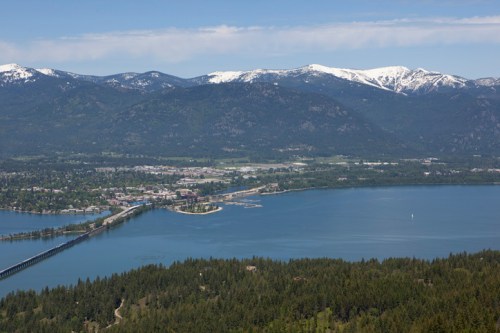 Aerial view of a lake surrounded by mountains, with a town and a bridge visible in the foreground.