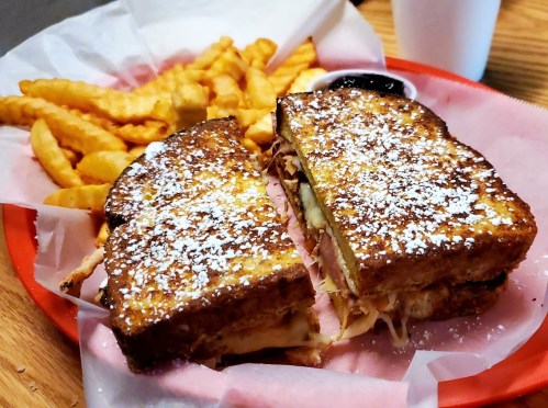 A plate with a sliced sandwich dusted with powdered sugar, served with crinkle fries and a small cup of sauce.