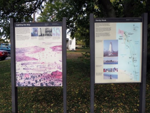 Two informational signs in a park, one about Sandy Hook and the other titled "Lighting the Way," with historical images and maps.