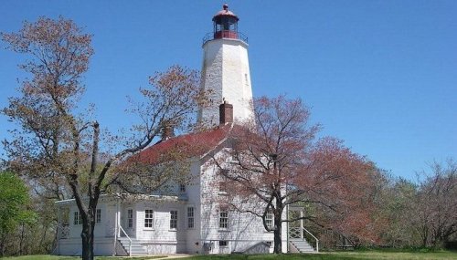 A white lighthouse with a red roof stands against a clear blue sky, surrounded by trees in early spring.