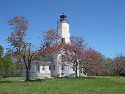 A white lighthouse with a red roof stands beside trees on a grassy area under a clear blue sky.