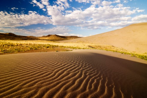 A serene desert landscape with rippled sand dunes under a blue sky dotted with clouds.