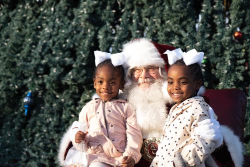 Two smiling girls with bows in their hair sit on Santa's lap in front of a decorated Christmas tree.