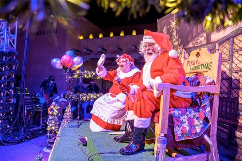 Santa and Mrs. Claus wave from their festive stage, surrounded by Christmas decorations and colorful balloons.