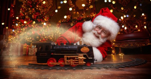 Santa lays under the Christmas tree with a model train car passing through with magical lights surrounding them.