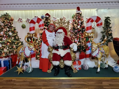 A festive scene with Santa Claus and Mrs. Claus surrounded by Christmas trees, candy canes, and reindeer decorations.
