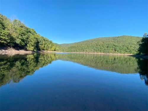 A calm lake surrounded by lush green trees and rolling hills under a clear blue sky. Reflections on the water's surface.