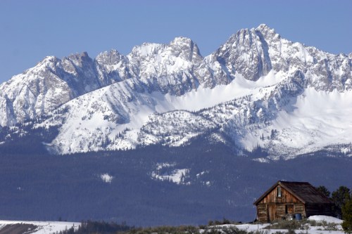 A rustic wooden cabin in the foreground with snow-covered mountains in the background under a clear blue sky.