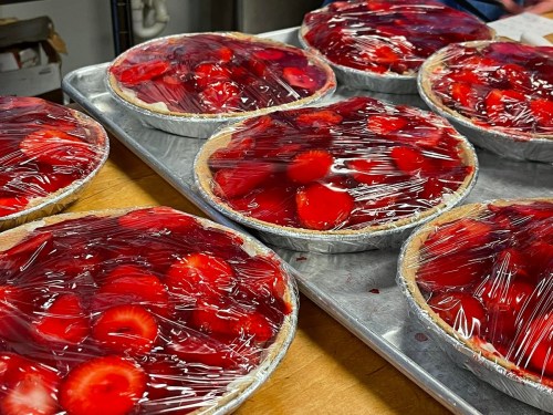 Six strawberry pies covered in plastic wrap on a baking tray, with vibrant red strawberries visible on top.
