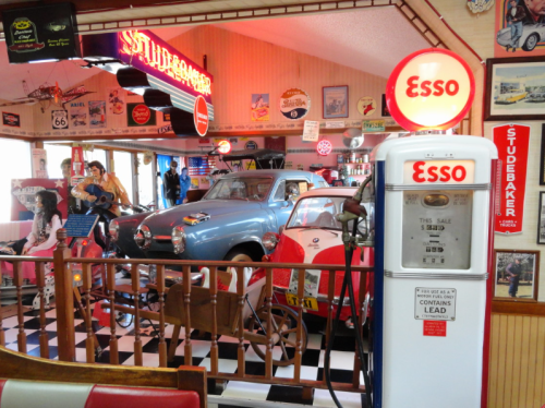 A vintage diner filled with classic cars, retro signs, and an old Esso gas pump, creating a nostalgic atmosphere.