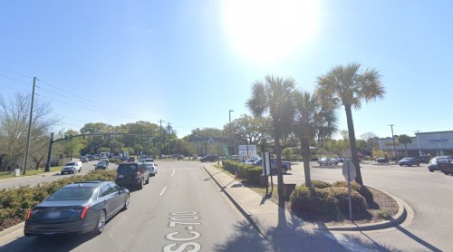 A sunny street scene with palm trees, cars, and shops along the road. Clear blue sky in the background.