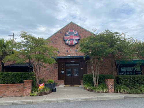 Exterior of a brick restaurant with a colorful sign, surrounded by greenery and a cloudy sky.