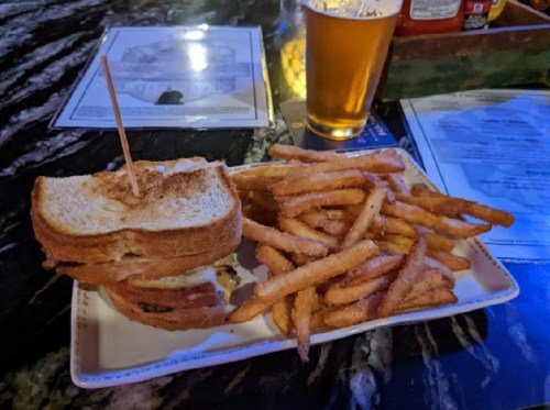A plate with a sandwich and a side of crispy fries, accompanied by a glass of beer in a dimly lit setting.