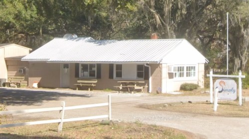 A single-story building with a metal roof, surrounded by trees, featuring picnic tables outside and a sign that reads "Simply."