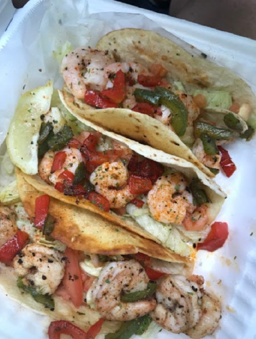 Three shrimp tacos filled with lettuce, tomatoes, and green peppers, served with lime wedges in a takeout container.