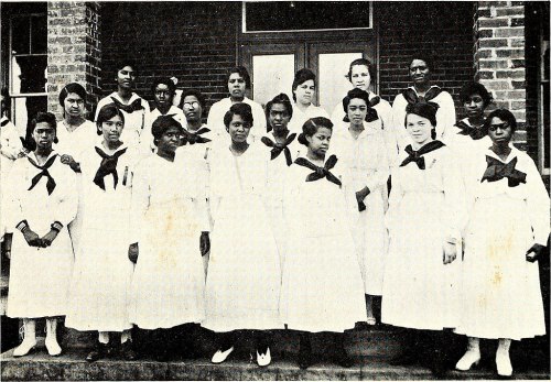 A group of young women in white dresses with dark bows, posing together outside a brick building.