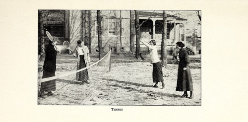 Four women playing tennis outdoors, dressed in early 20th-century attire, with a house and trees in the background.