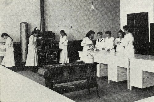 A historical classroom scene with women in white aprons engaged in various activities at tables and a stove.