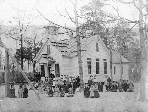 A historic black-and-white photo of a schoolhouse with children and adults gathered outside, surrounded by trees.
