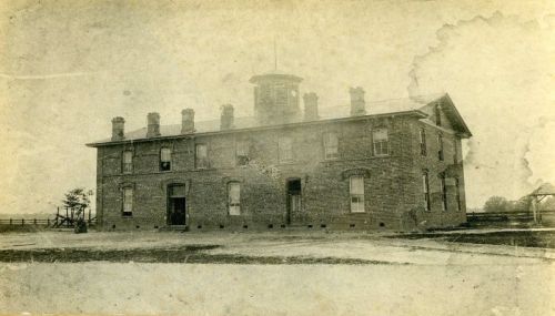 Historic brick building with multiple windows and a clock tower, set in a rural landscape. Black and white photo.