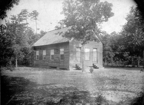 A vintage black-and-white photo of a wooden house surrounded by trees, featuring steps and multiple windows.